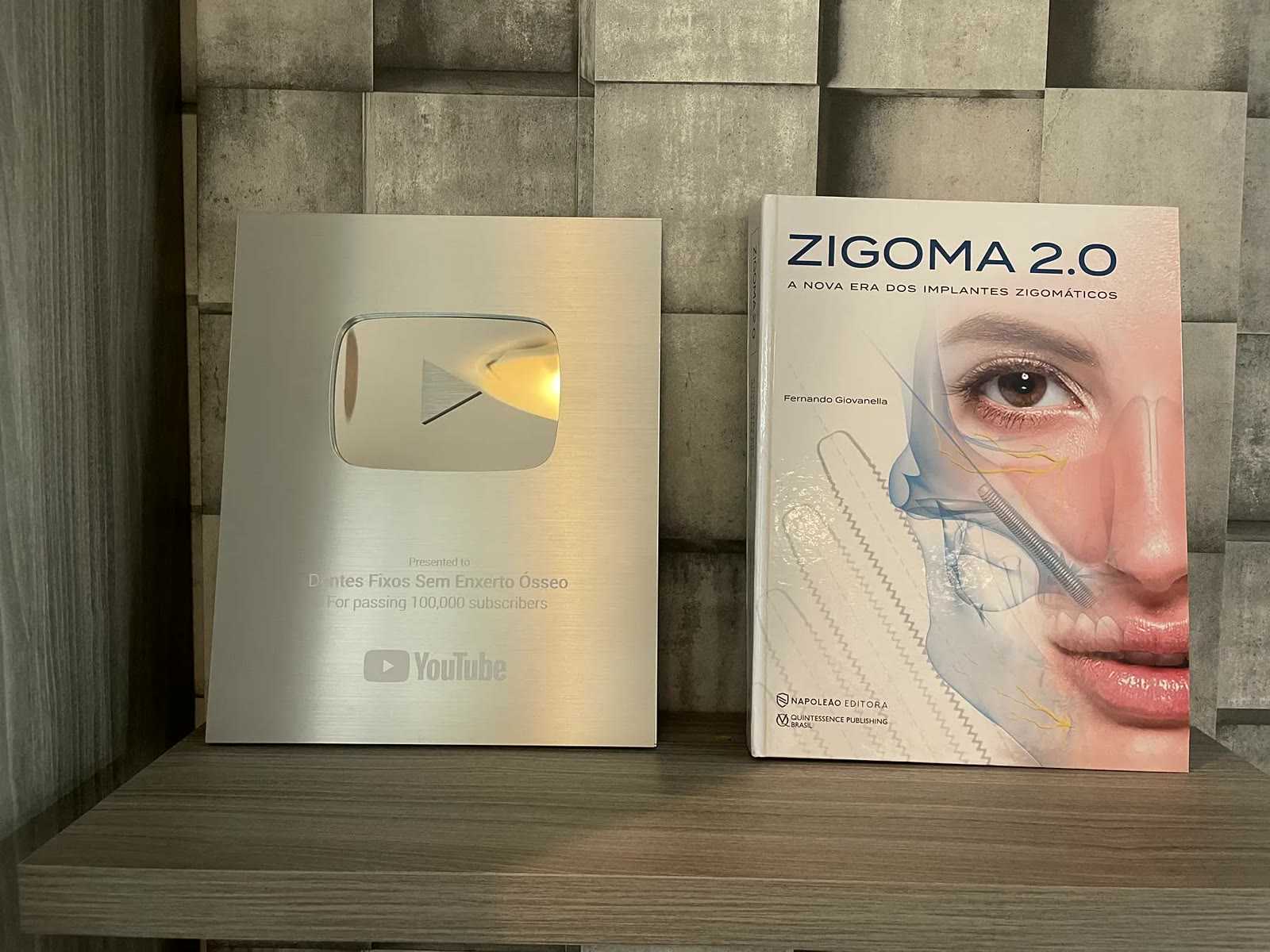 The book ZIGOMA 2.0 by Dr. Fernando Giovanella next to the YouTube plaque for 100,000 subscribers